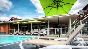 A poolside area with lounge chairs and umbrellas, a clear sky, and a building with a tiled roof in the background.