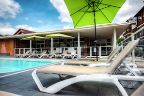 Zona junto a la piscina con sillones de descanso y sombrillas, cielo despejado y un edificio con tejado de tejas al fondo.