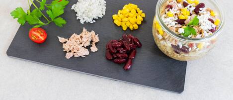 A variety of food items including tomatoes, rice, corn, tuna, and kidney beans arranged on a slate board, with a jar of mixed salad visible.