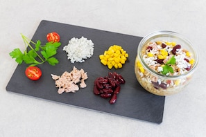 Une variété d’aliments, notamment des tomates, du riz, du maïs, du thon et des haricots rouges, sont disposés sur une planche d’ardoise, et un bocal de salade composée est visible.