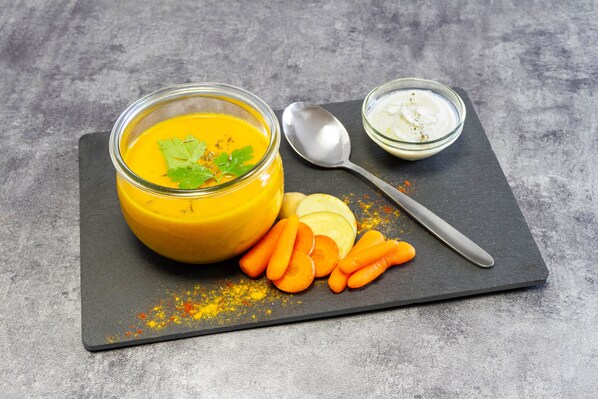 Un tazón de sopa amarilla con adorno, una cuchara y un pequeño recipiente de salsa blanca sobre una tabla de pizarra con palitos de zanahoria.
