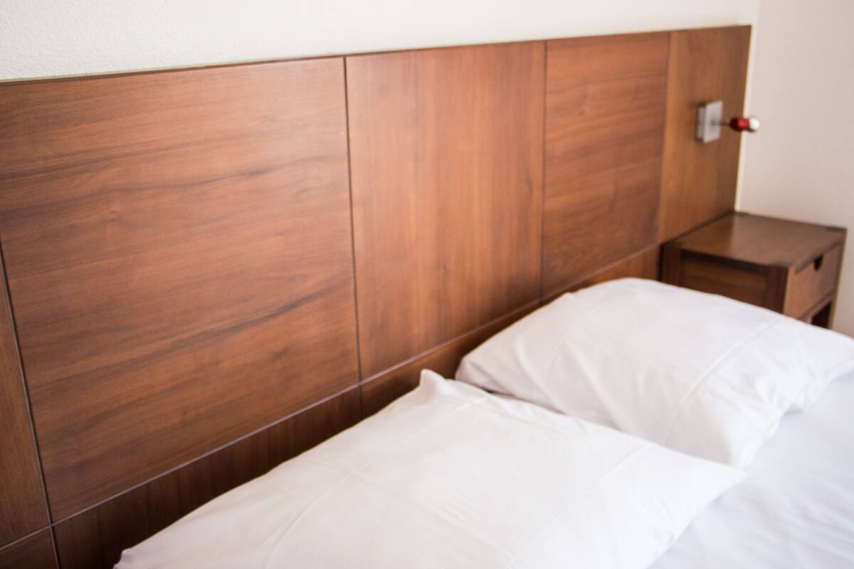 Una cama bien hecha con sábanas blancas, un cabecero de madera y una mesita de noche con un objeto rojo encima.