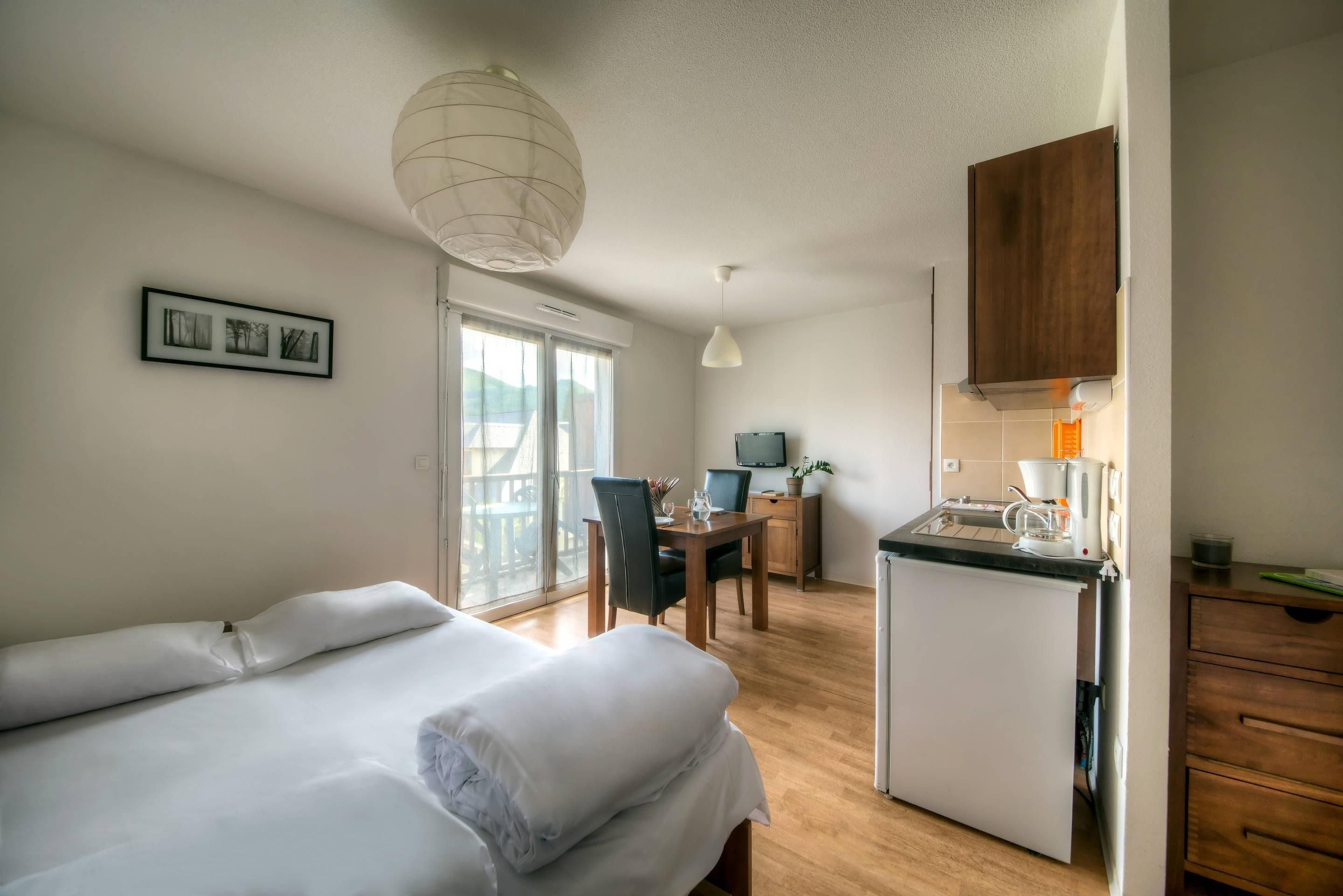 A hotel room with a bed, a desk with a chair, a kitchenette, and a large window.