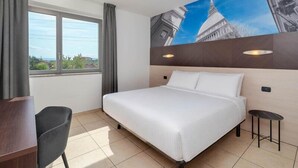A hotel room with a large bed, a desk, a chair, a window with curtains, and a wall with a cityscape mural.
