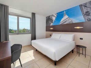 A hotel room with a large bed, a desk, a chair, a window with curtains, and a wall with a cityscape mural. - B&B Hotel Settimo Torinese (Settimo Torinese)