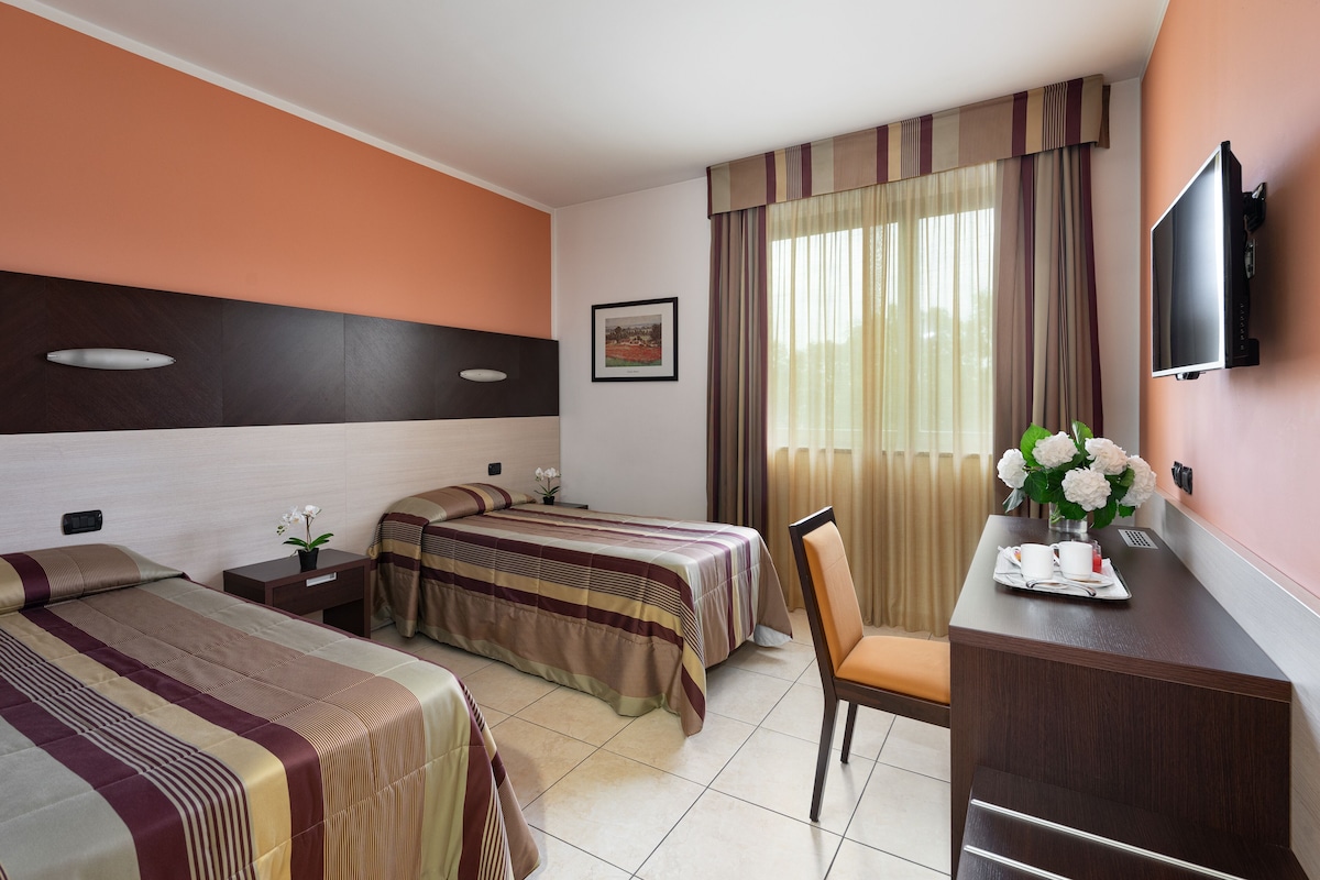 Una habitación de hotel con dos camas, un escritorio con televisor y una ventana con cortinas.