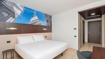 A hotel room with a large bed, a small table, a chair, a wall-mounted artwork, and a door.