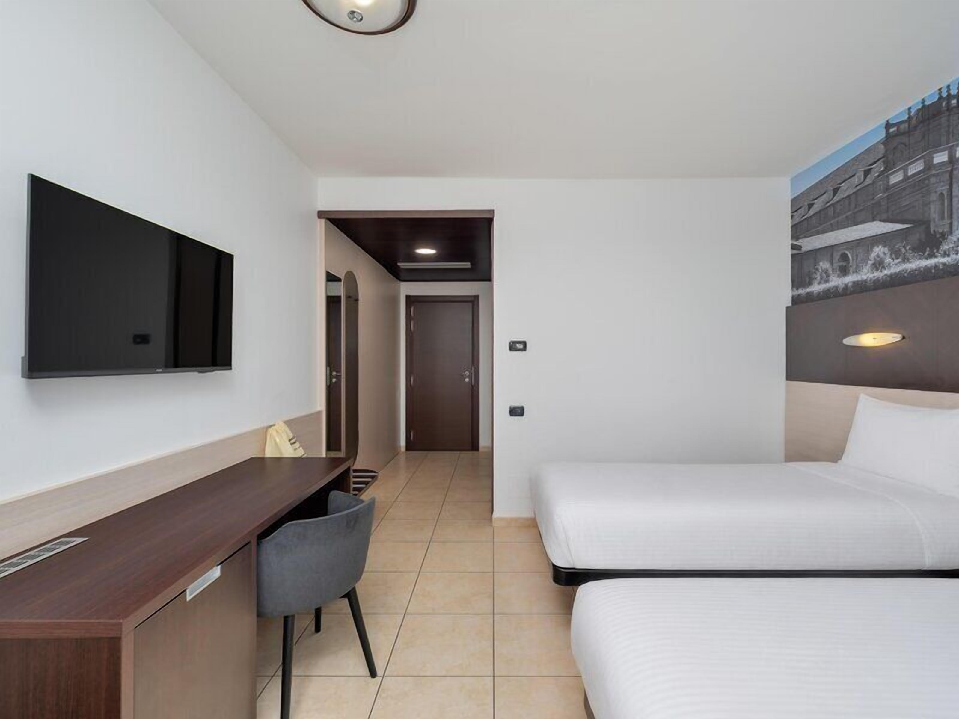 Quarto de hotel com uma cama, uma escrivaninha, uma cadeira, uma televisão fixada na parede e uma porta que dá para outro cômodo.