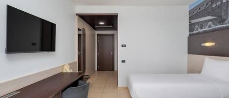 A hotel room with a bed, a desk, a chair, a TV mounted on the wall, and a door leading to another room.