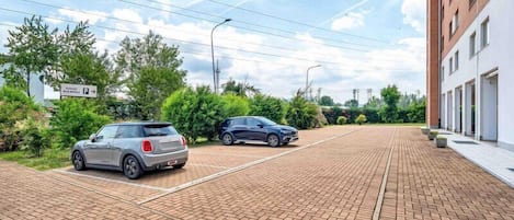 A parking area with two cars, a brick pathway, and a building with multiple windows.