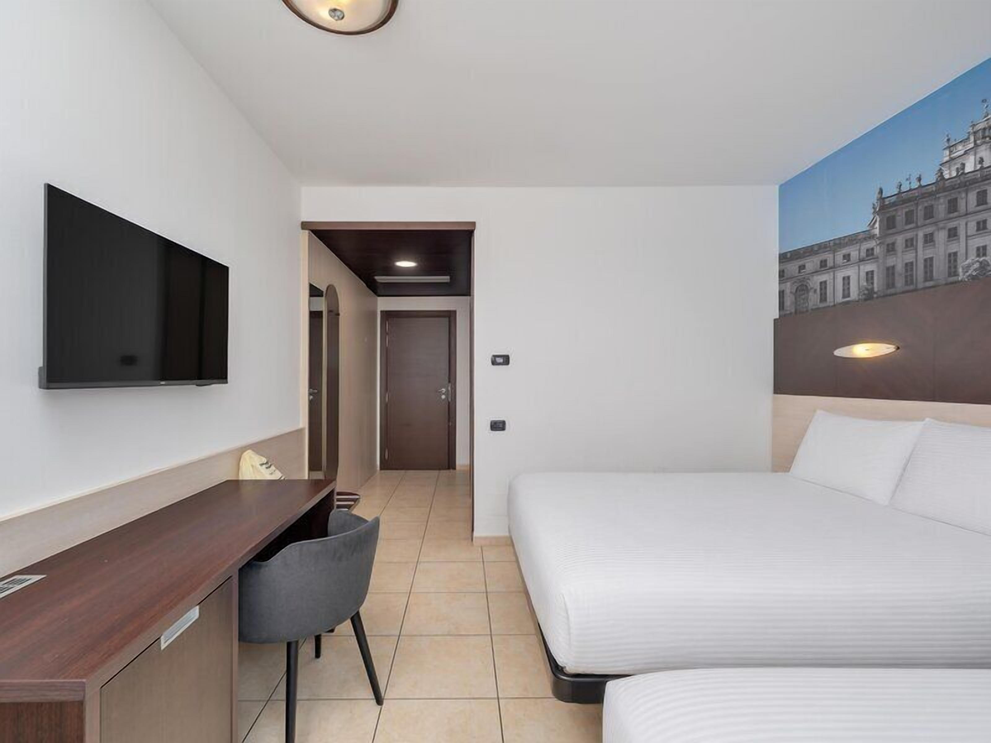 A hotel room with a bed, a desk, a chair, a TV, and a painting on the wall.