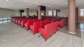 A large indoor space with rows of red chairs, a flat ceiling with recessed lighting, and a polished floor.