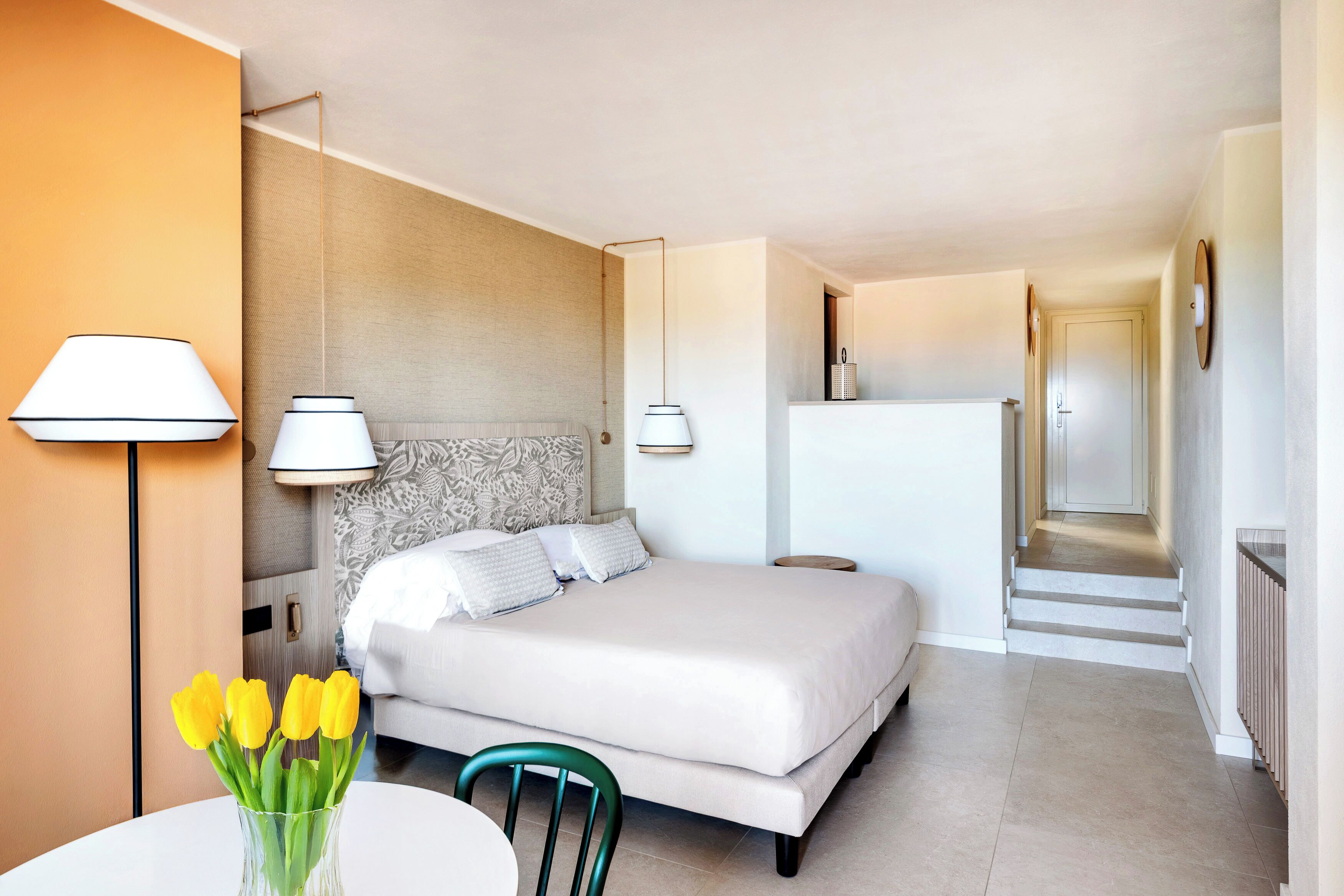 A hotel room with a bed, bedside lamps, a vase with flowers, a chair, and a table.