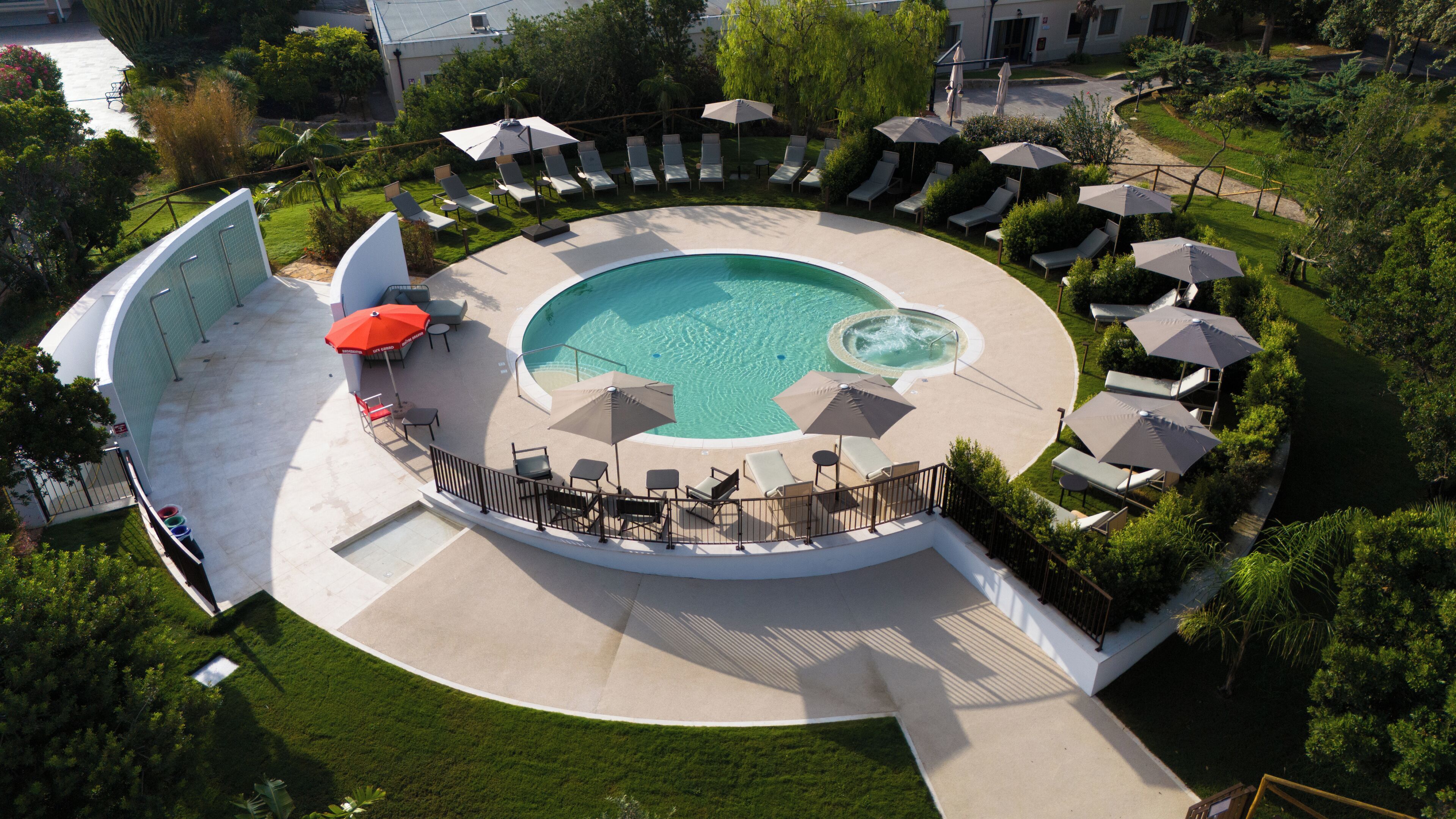 Una piscina circular con sillas de descanso y sombrillas, rodeada por un césped cuidado y árboles.