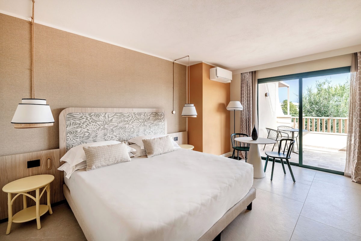 A hotel room with a large bed, a small table, a chair, and a balcony with outdoor seating.
