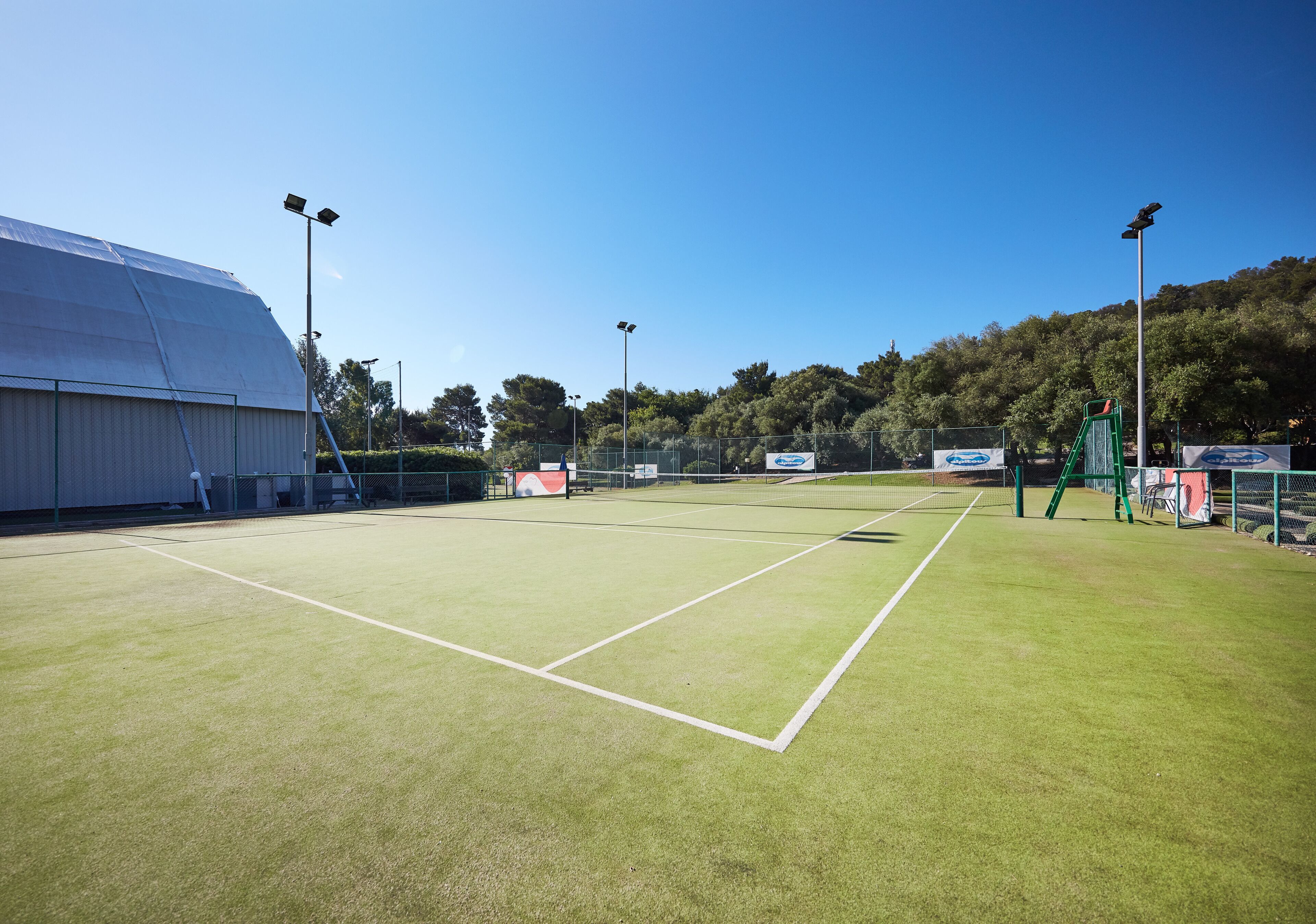 Una cancha de tenis con una red, rodeada por una valla, y un edificio grande a la izquierda.