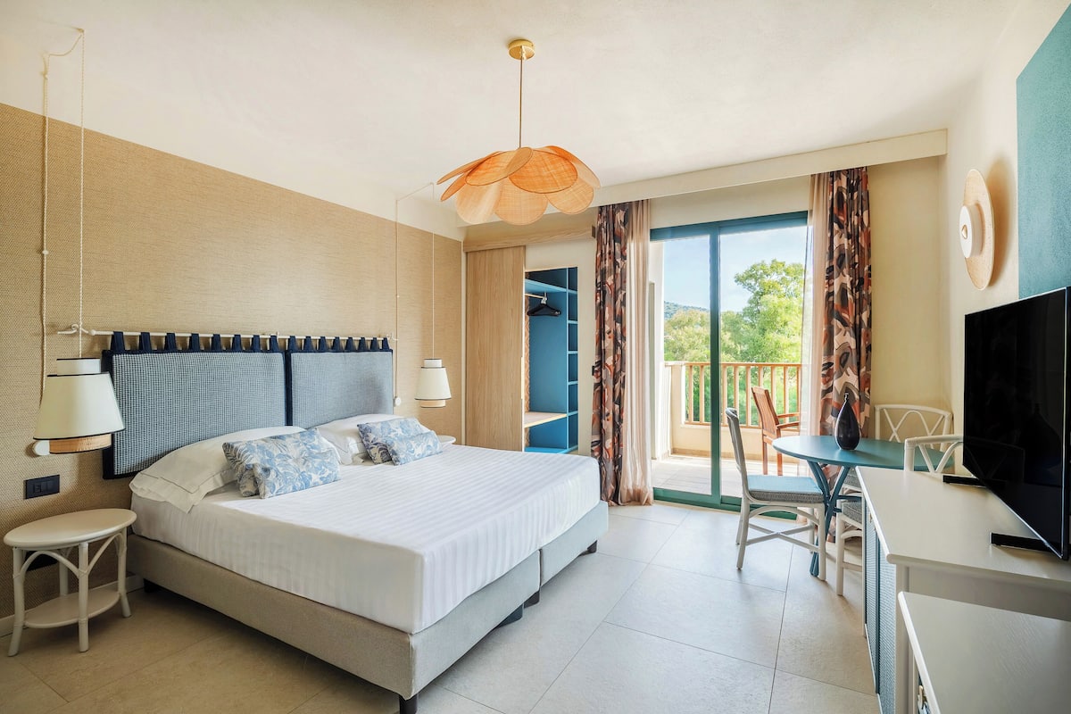 A hotel room with a bed, bedside tables, a desk, a television, and a balcony with a view.