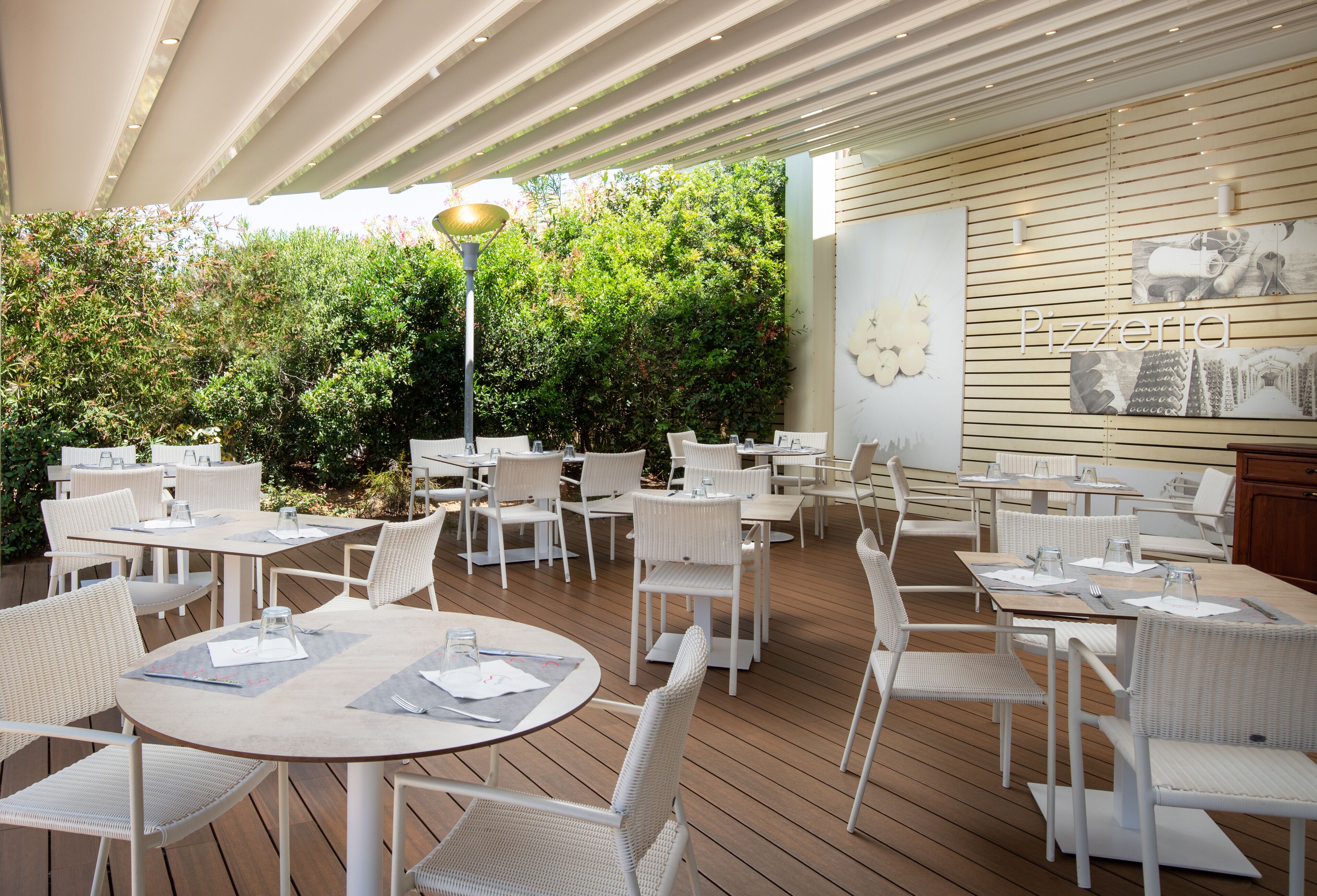 An outdoor dining area with white wicker furniture and tables, surrounded by greenery.
