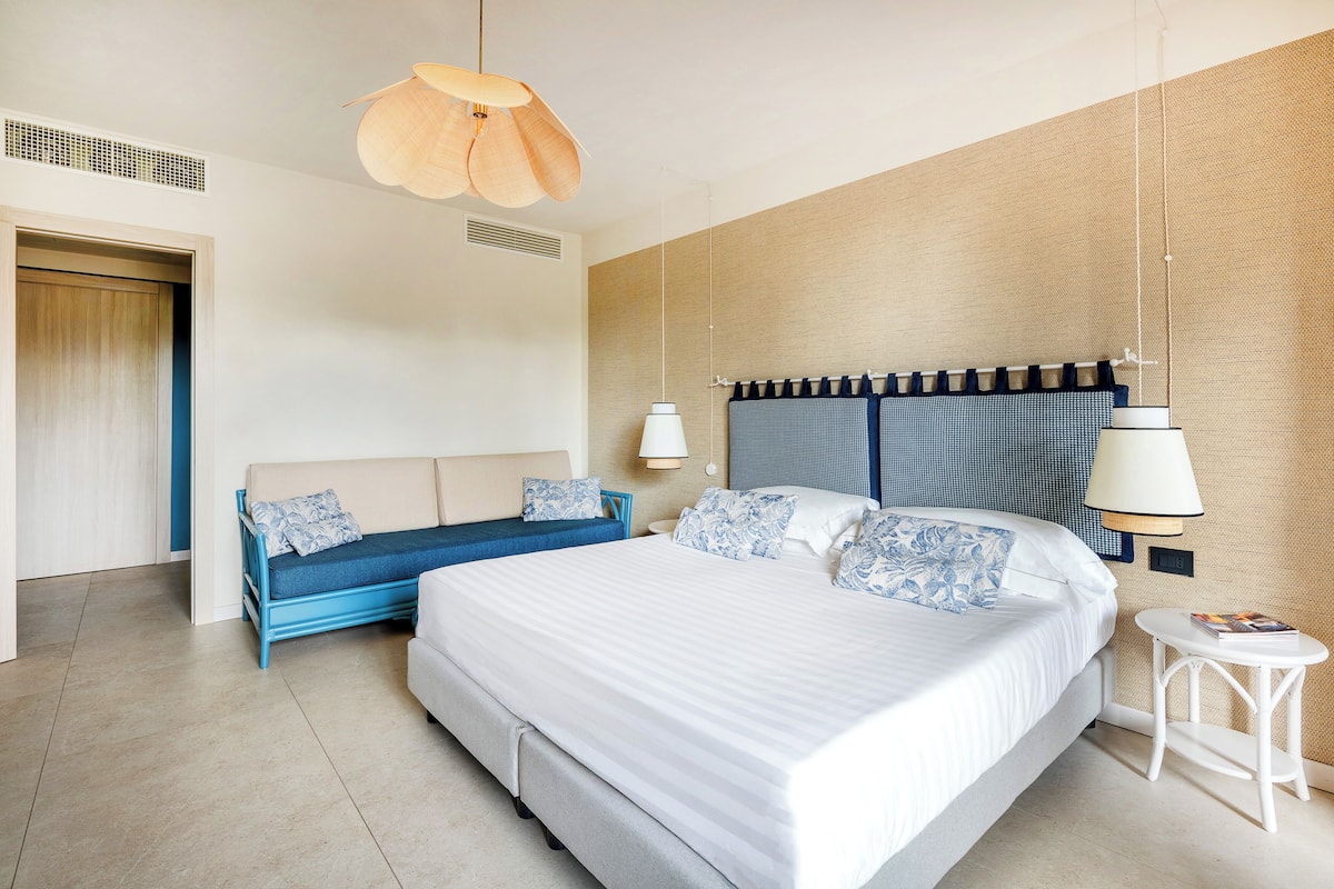 A modern hotel room with a large bed, a blue sofa, a small table, and a hanging light fixture.
