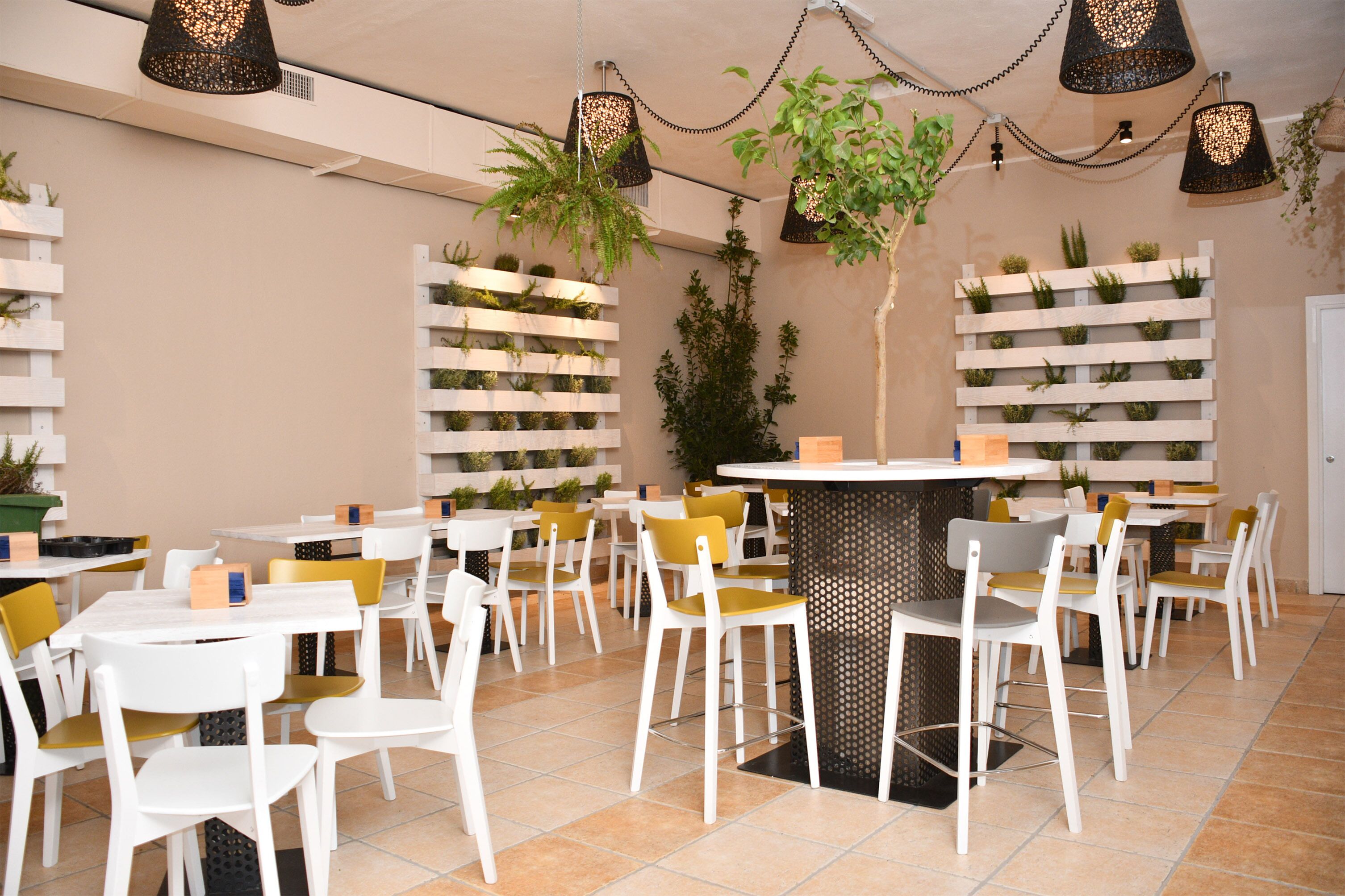 Ein Restaurant mit weißen Tischen und Stühlen, hängenden Pflanzen und schwarzen Pendelleuchten.