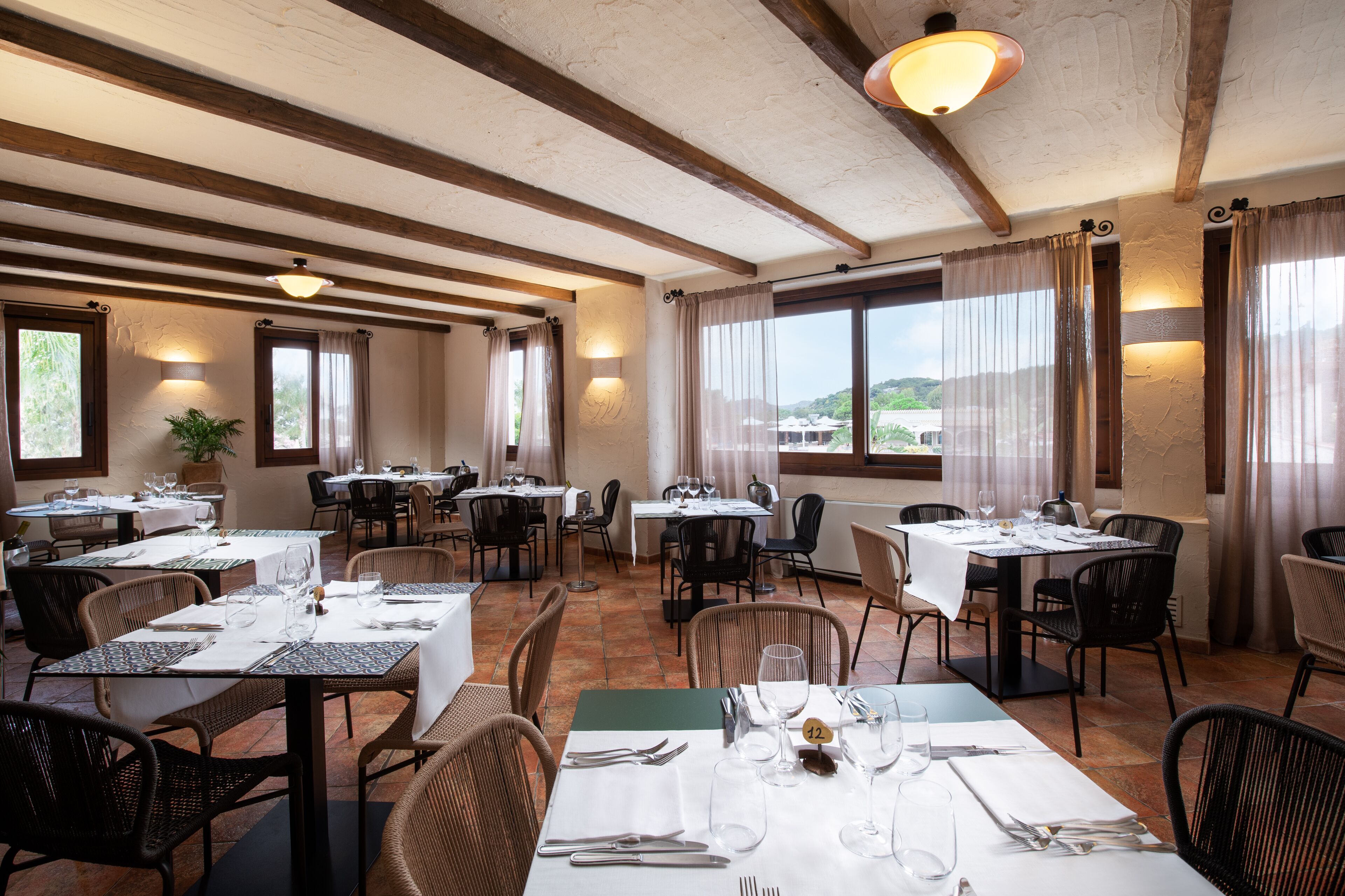Een restaurant met meerdere tafels en stoelen, houten balken in het plafond en grote ramen met gordijnen.