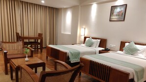 A hotel room with two beds, wooden furniture, a dining table, and a large window. - Grand Hotel (Kanayannur)