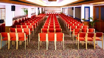 Un salón de conferencias con filas de sillas rojas, alfombra estampada y paneles de madera en las paredes.