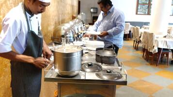 Un chef prepara comida en una cocina con equipo de acero inoxidable y un bufé al fondo.