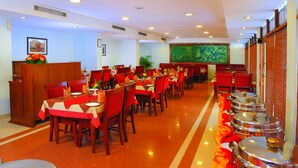 A restaurant with multiple dining tables and chairs, a buffet line with food containers, and a large decorative wall painting. - Hotel Excellency (Kanayannur)