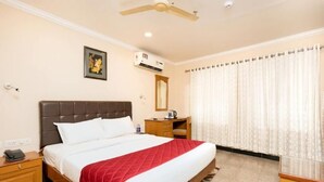 A hotel room with a bed, a desk, a chair, a ceiling fan, an air conditioning unit, and a window with curtains. - Hotel Excellency (Kanayannur)