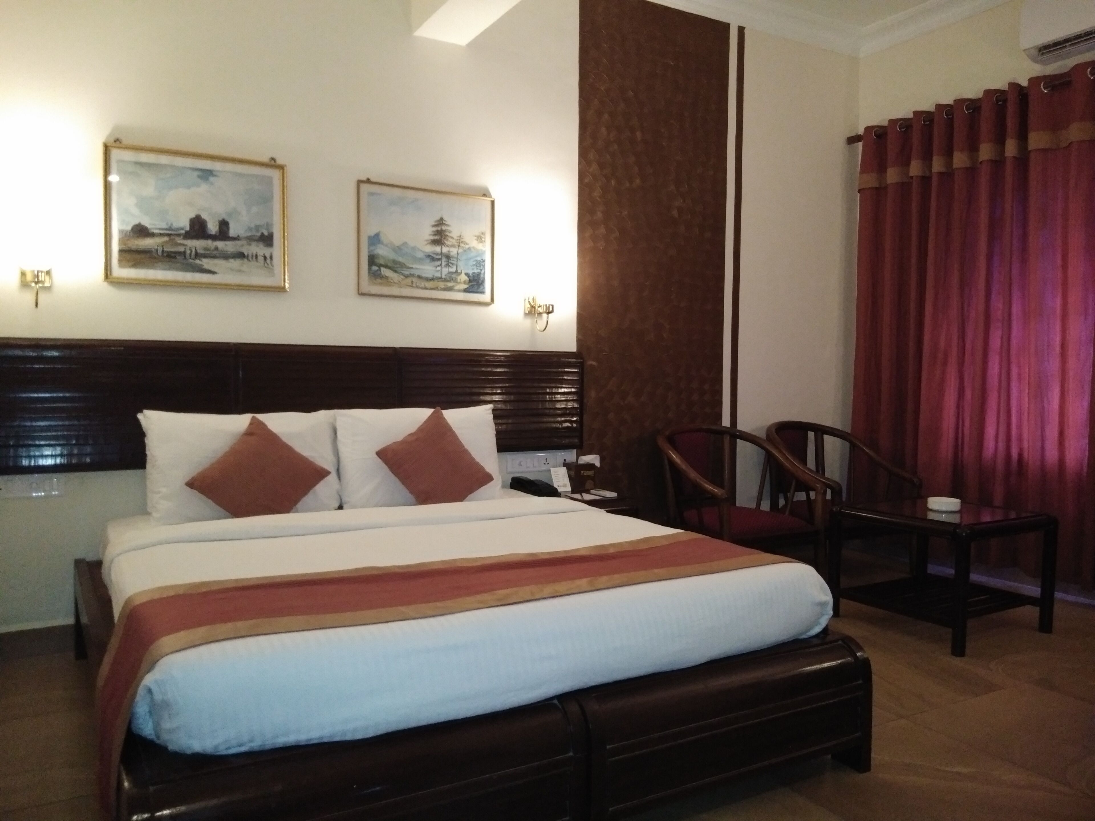 Une chambre d’hôtel avec un grand lit, deux tableaux accrochés au mur, une tête de lit en bois, un rideau rouge et une petite table avec une chaise.