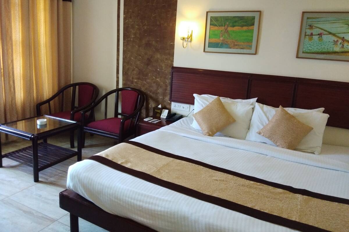 Quarto de hotel com uma cama, duas cadeiras, uma mesinha e duas pinturas na parede.
