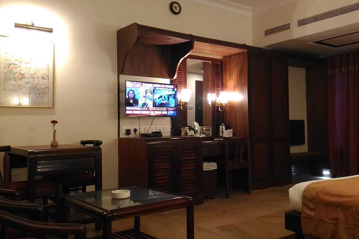 Quarto de hotel com cama, escrivaninha com cadeira, televisão e uma mesinha.