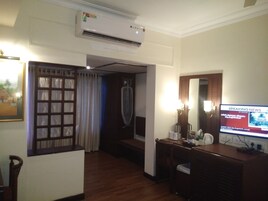 A hotel room with a wooden floor, a television displaying news, a wall-mounted air conditioner, and a large mirror.