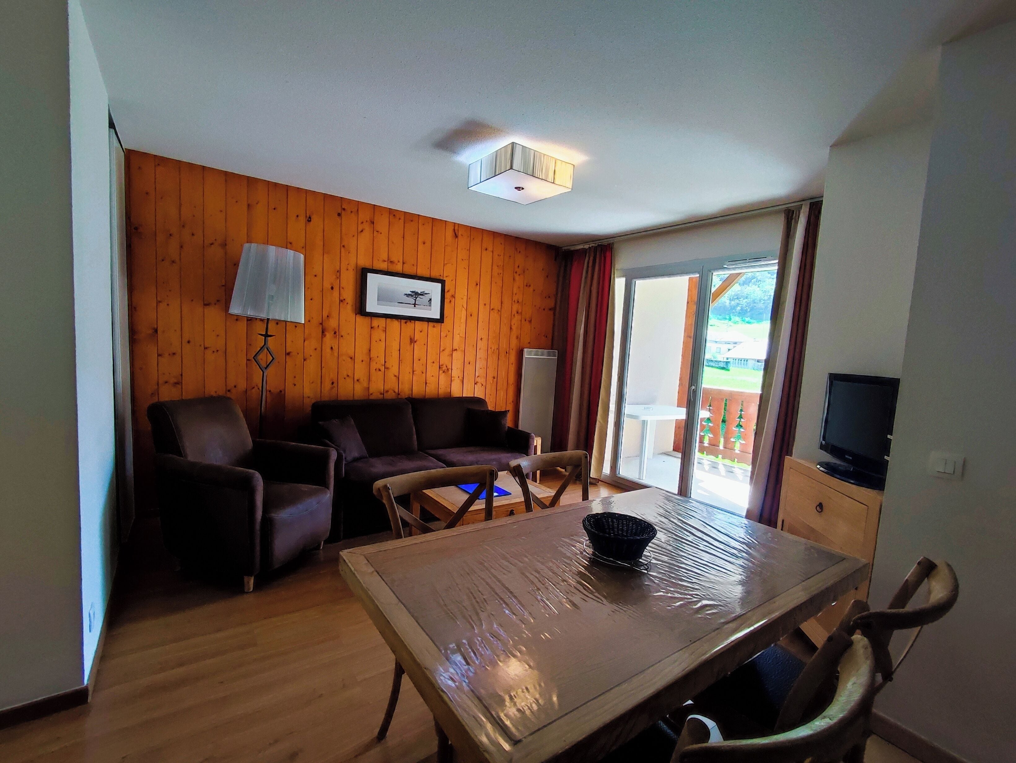 Habitación con piso y pared de madera, un sofá, una silla, una mesa con una cesta, un televisor y una puerta corrediza de vidrio que da a un área exterior con una mesa y sillas.