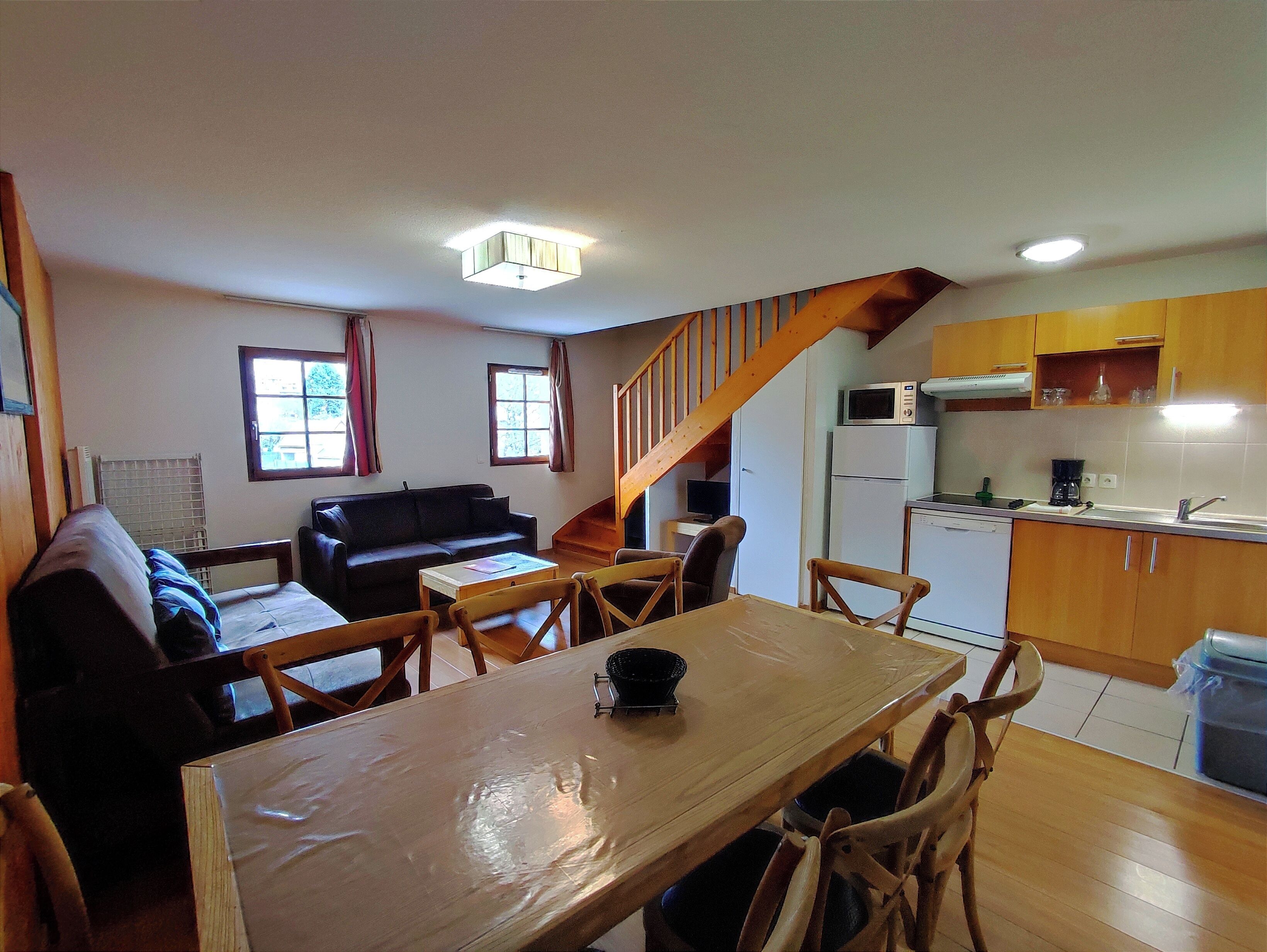 A compact living space with a dining table, wooden chairs, a sofa, a kitchen area with a microwave and refrigerator, and a staircase.