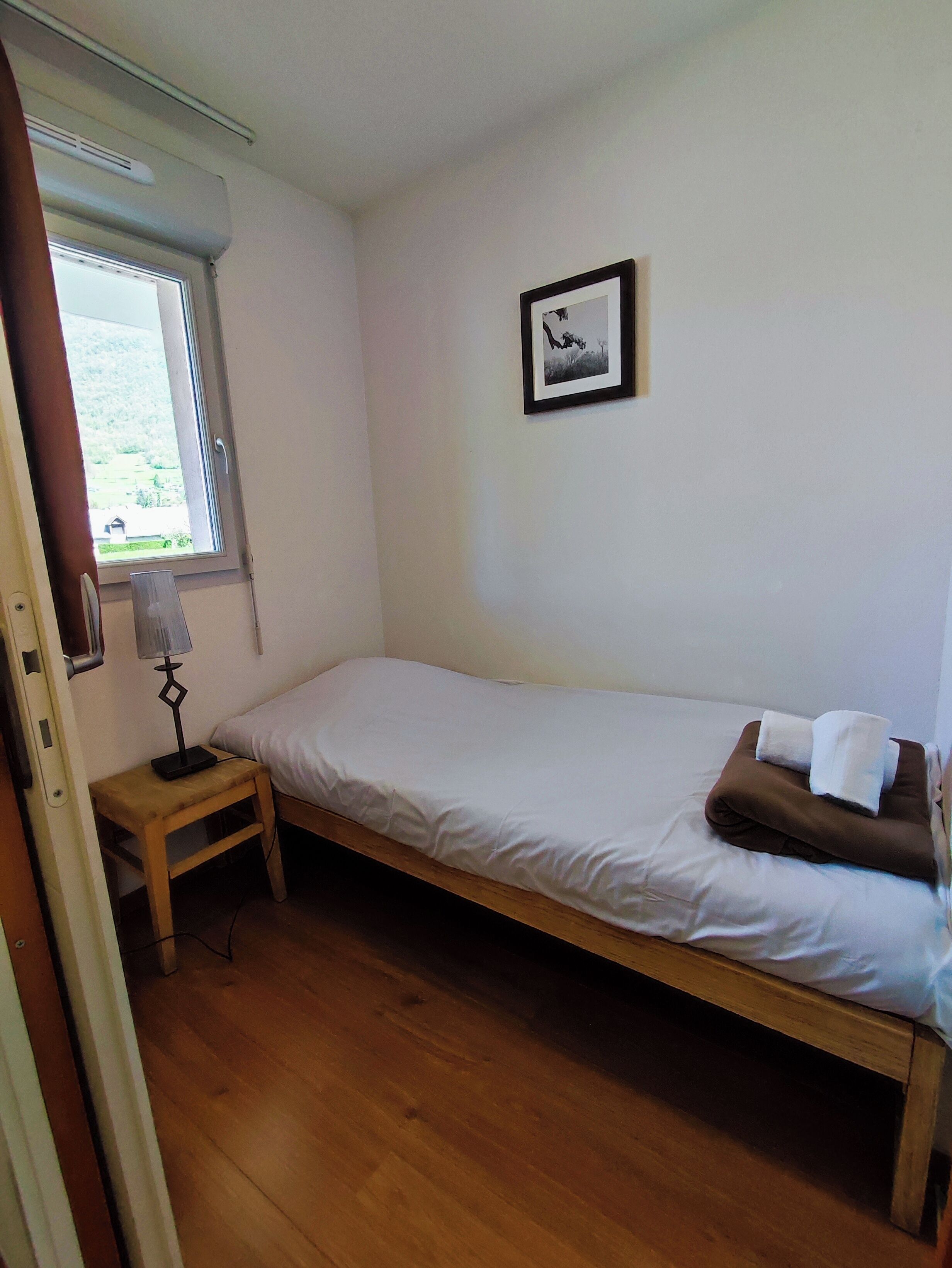 Quarto pequeno com uma cama de solteiro, piso de madeira, mesa de cabeceira com abajur, janela com vista para área verde e um quadro na parede.