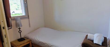 A small, single-bed room with a wooden floor, a bedside table with a lamp, a window with a view of greenery, and a framed picture on the wall.