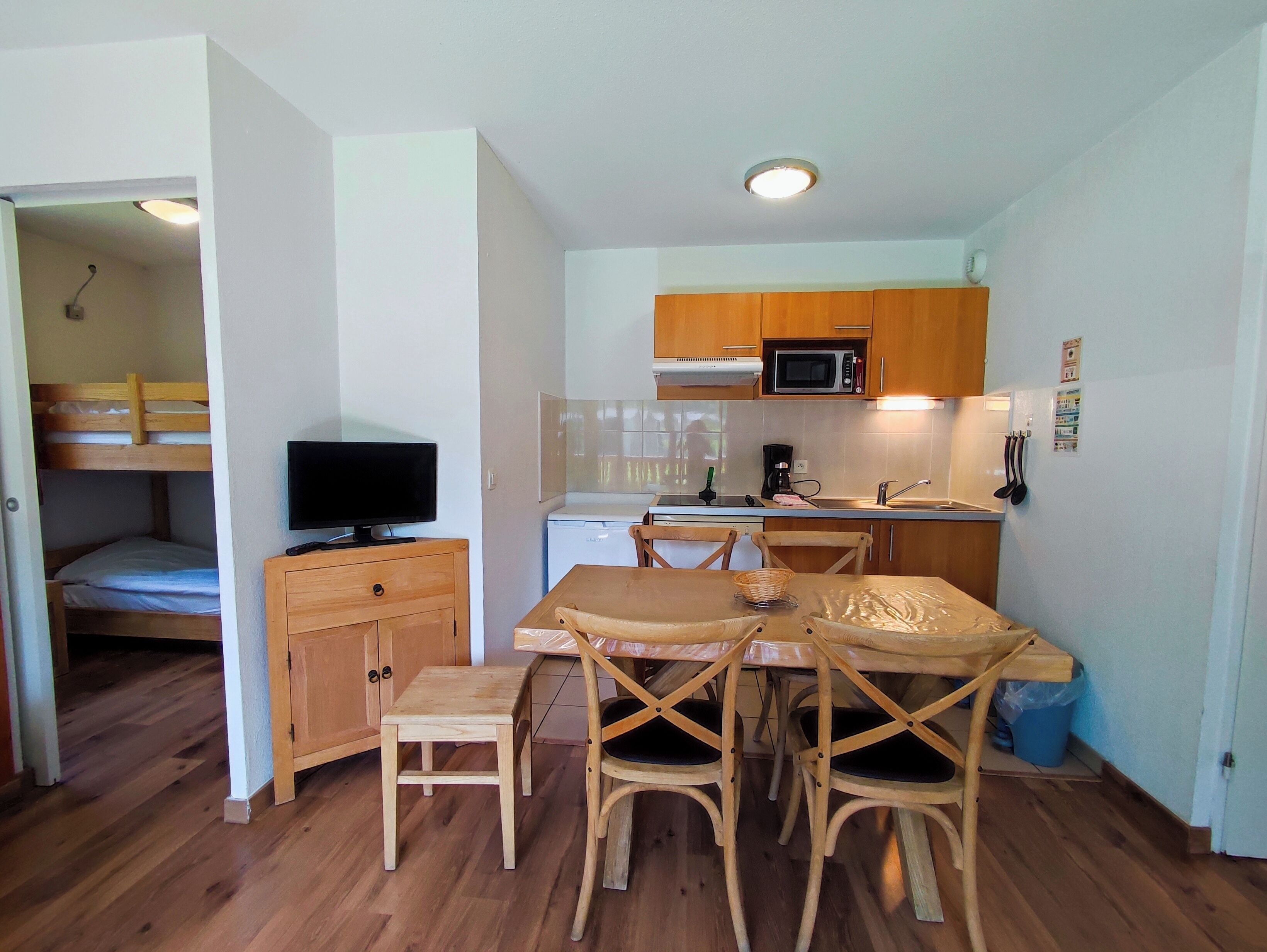A compact kitchen with wooden cabinets, a microwave, and a sink. There is a dining table with chairs and a small wooden stool. A television is placed on a wooden cabinet.