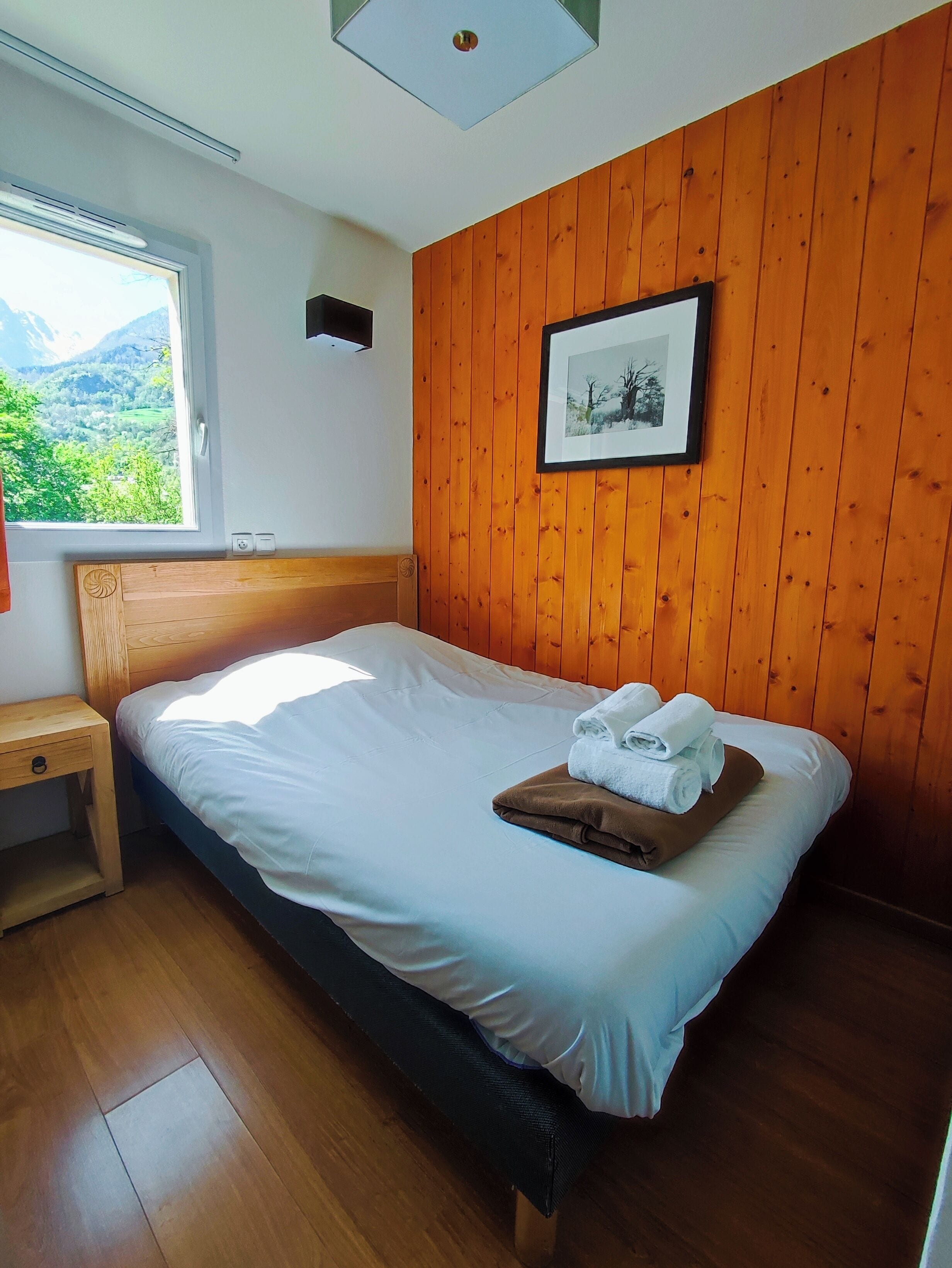 A bedroom with a wooden headboard, a bed with white bedding, a wooden nightstand, a window with a view of greenery, and a framed picture on the wall.