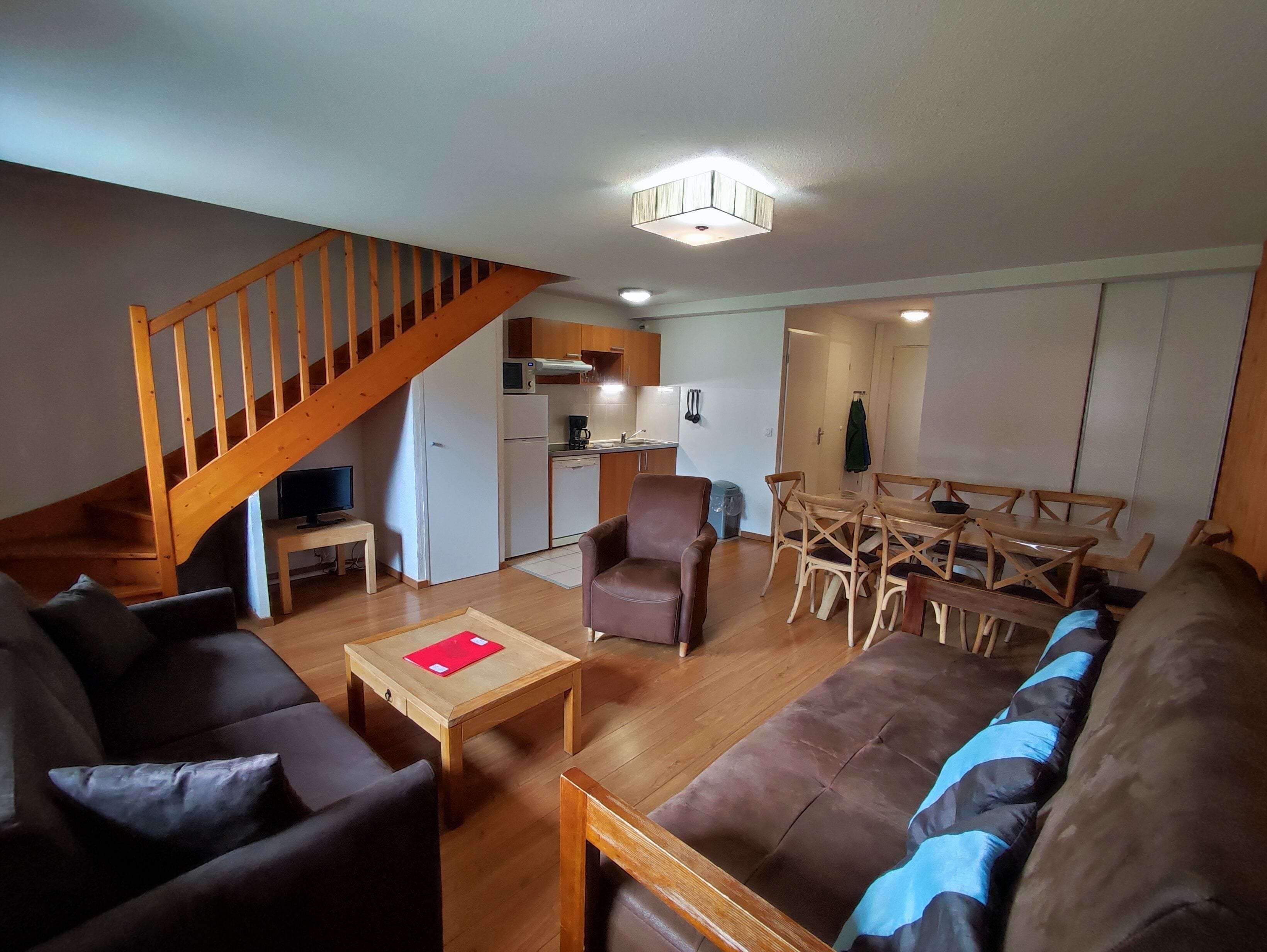 Uma sala de estar com sofá, poltrona, mesa de centro de madeira e um espaço de jantar com mesa e cadeiras. Uma escada é visível ao fundo.