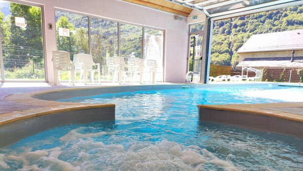 A hot tub with water jets, surrounded by a pool, with chairs and a view of a forested hillside. - Domaine Val de Roland (Luz-Saint-Sauveur)
