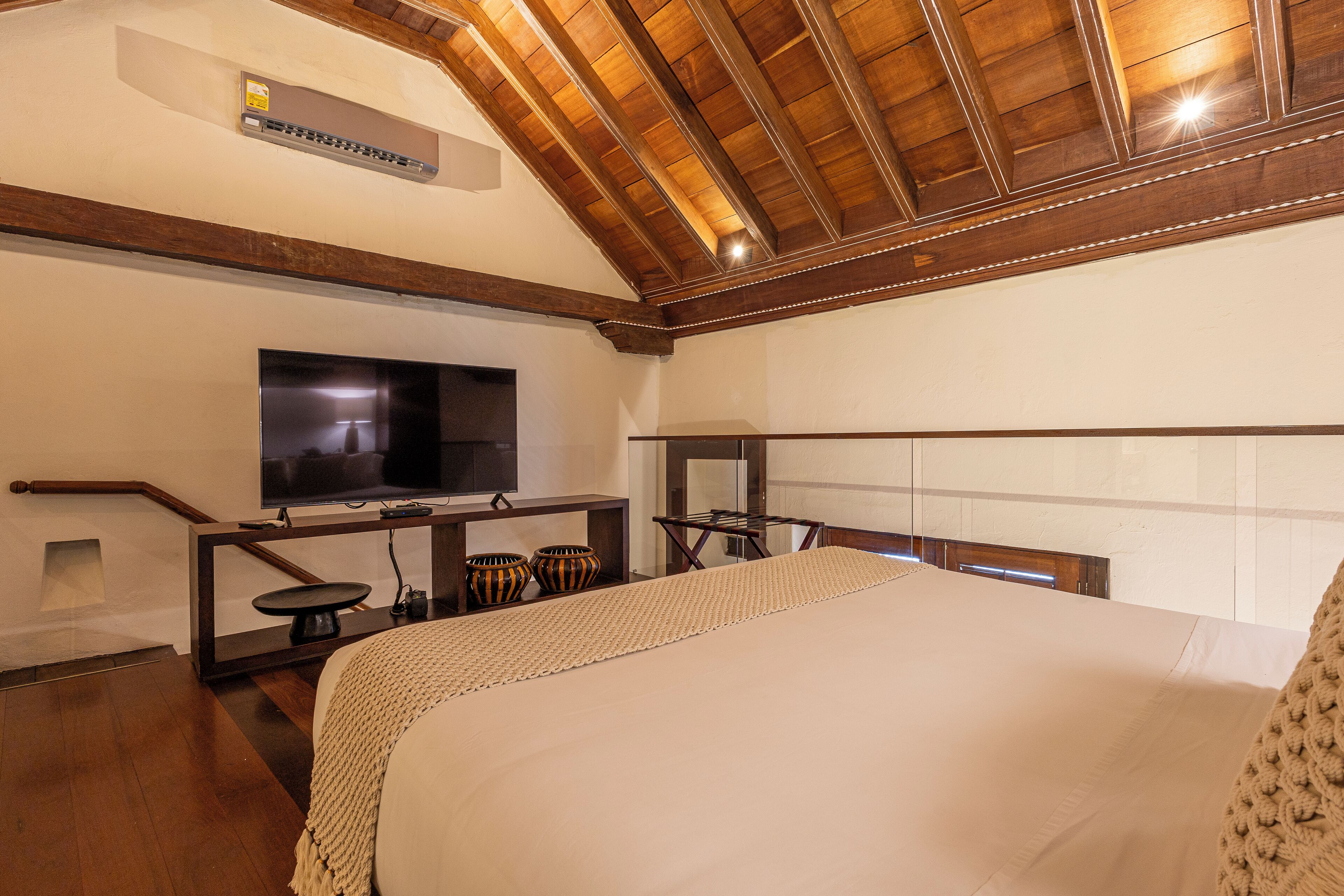 A hotel room with a large bed, a flat-screen TV mounted on the wall, wooden beams on the ceiling, and a small table with decorative bowls.