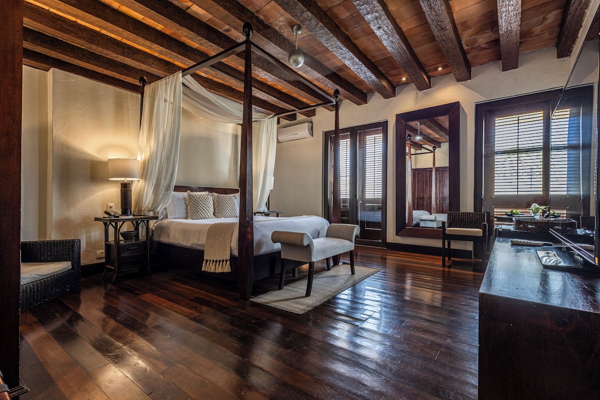 A spacious bedroom with a four-poster bed, a sitting area with a chair and a small table, and a large mirror reflecting the room.