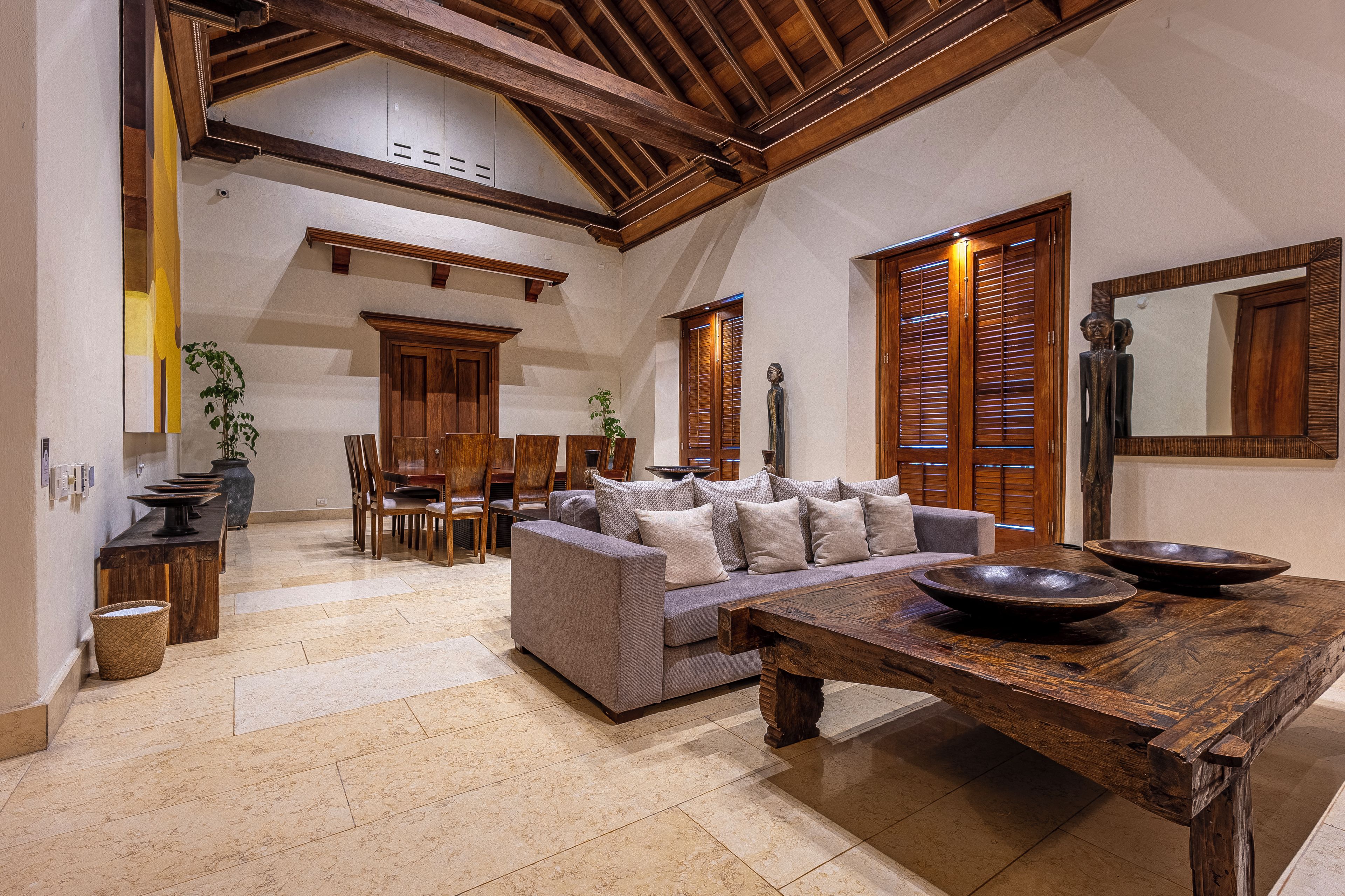 A spacious living area with a high ceiling, wooden beams, a sofa set, a dining table, and a large mirror.