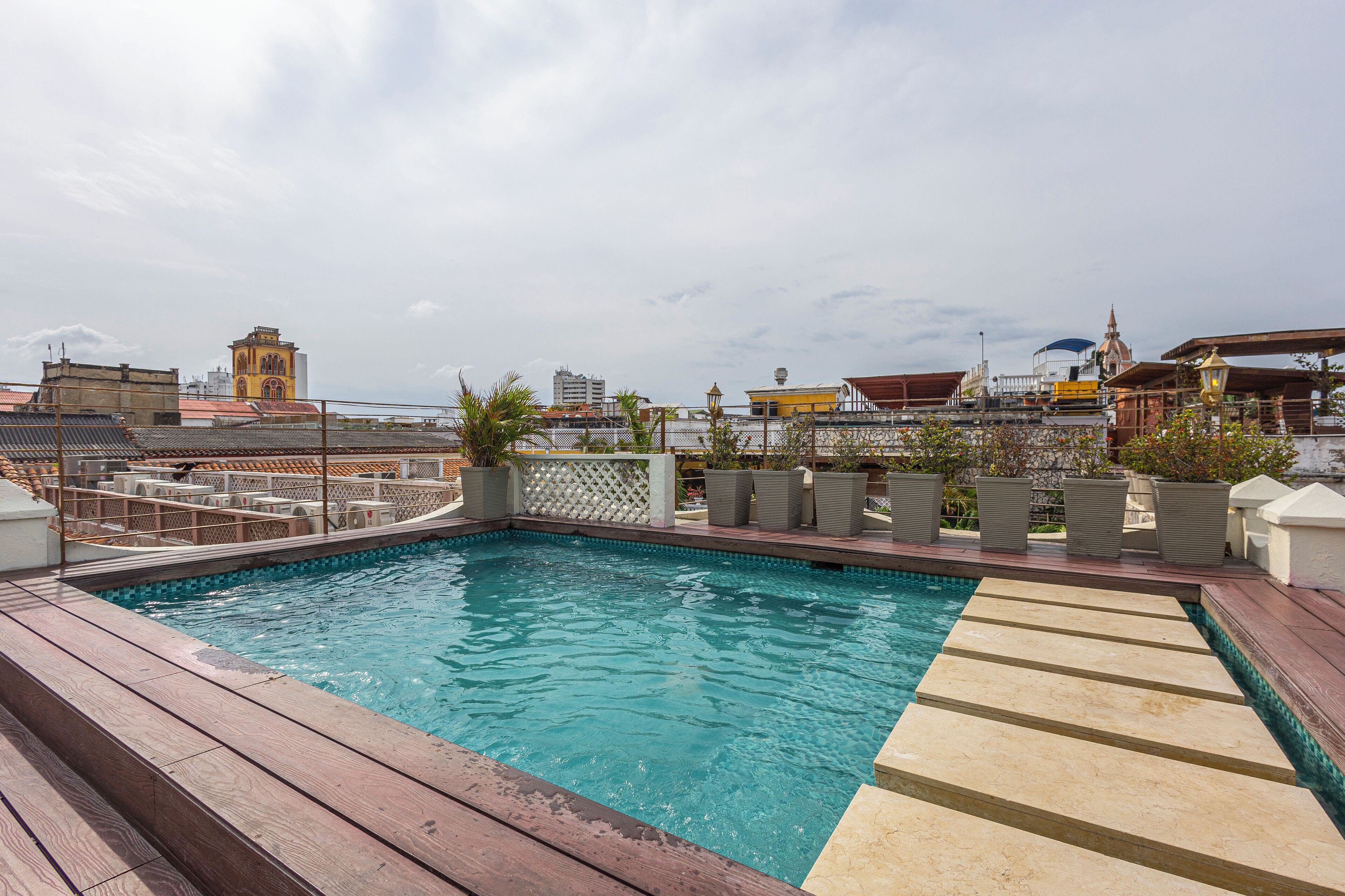A rooftop swimming pool with a wooden deck, surrounded by potted plants and a fence.