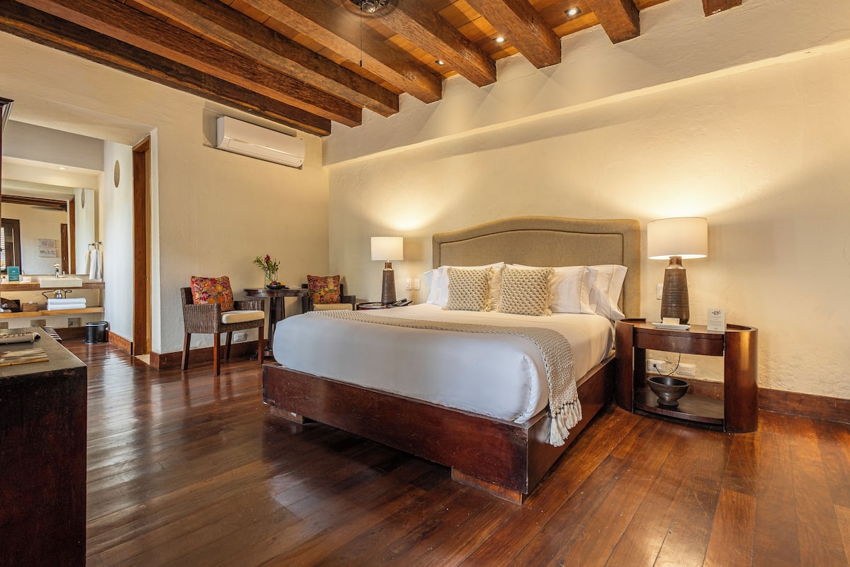 A spacious bedroom with a large bed, wooden beams on the ceiling, a bedside table with a lamp, and a small side table with a bowl.