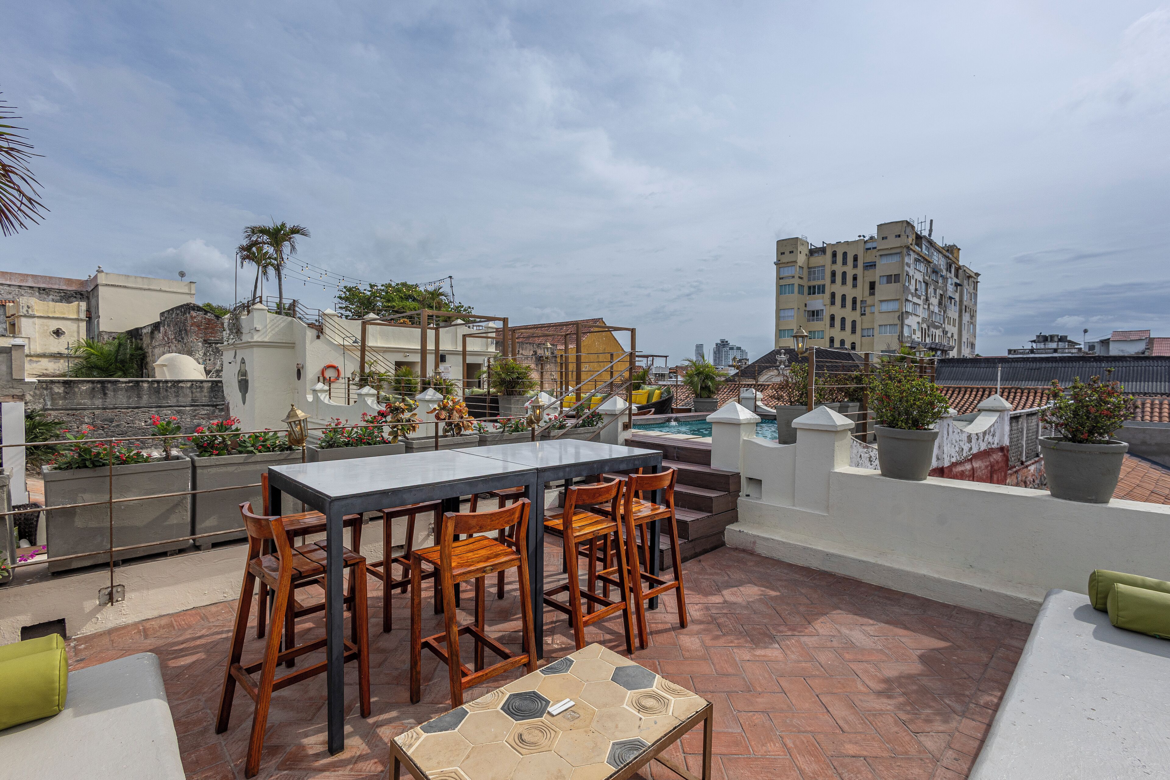 A rooftop terrace with a dining table and chairs, surrounded by potted plants and a swimming pool.