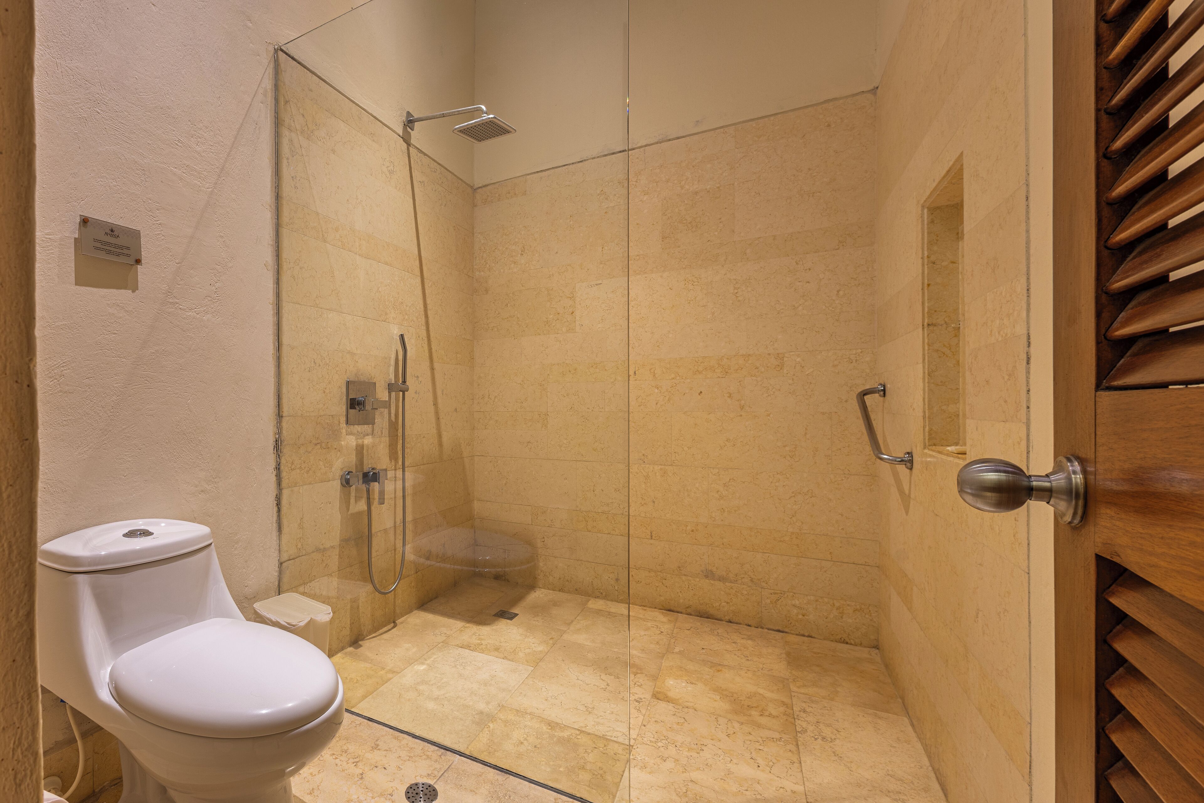 Un petit espace douche fermé, doté d’une porte en verre, de toilettes et d’une porte en bois avec une poignée.