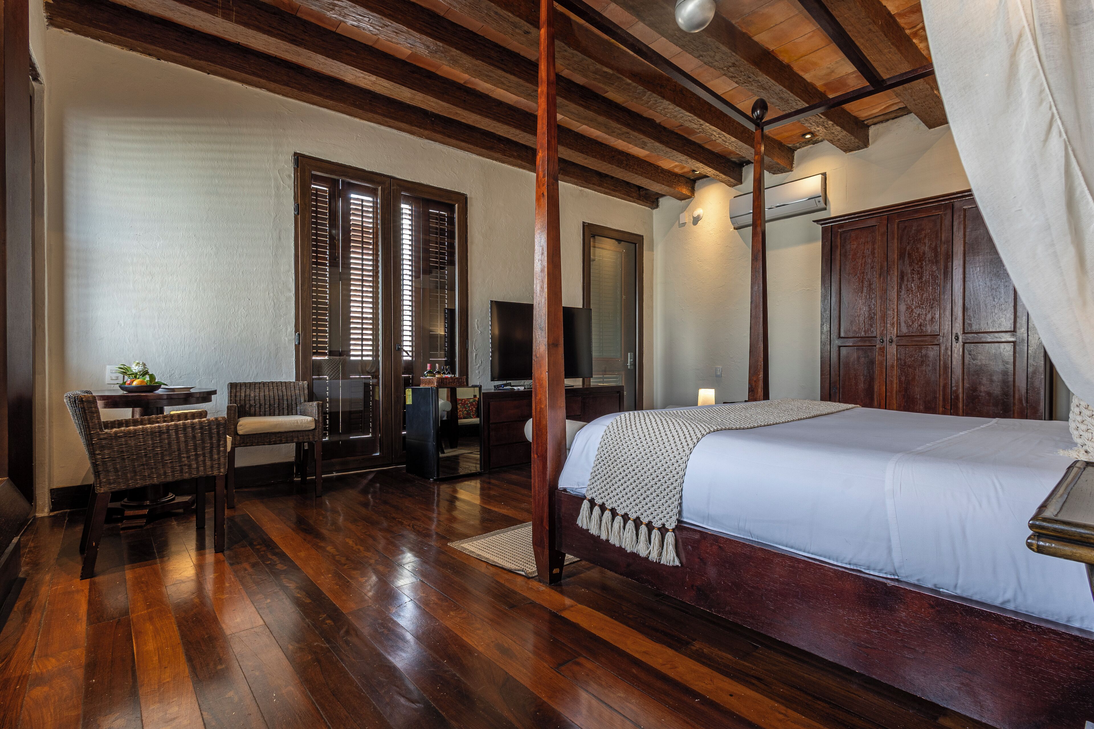 Une chambre à coucher avec un lit à baldaquin en bois, un coin salon avec des fauteuils en osier, une télévision et une armoire.